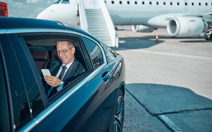 Cheerful businessman with smartphone in car at airport Smiling elegant man in glasses is using cell phone during transfer after trip by plane